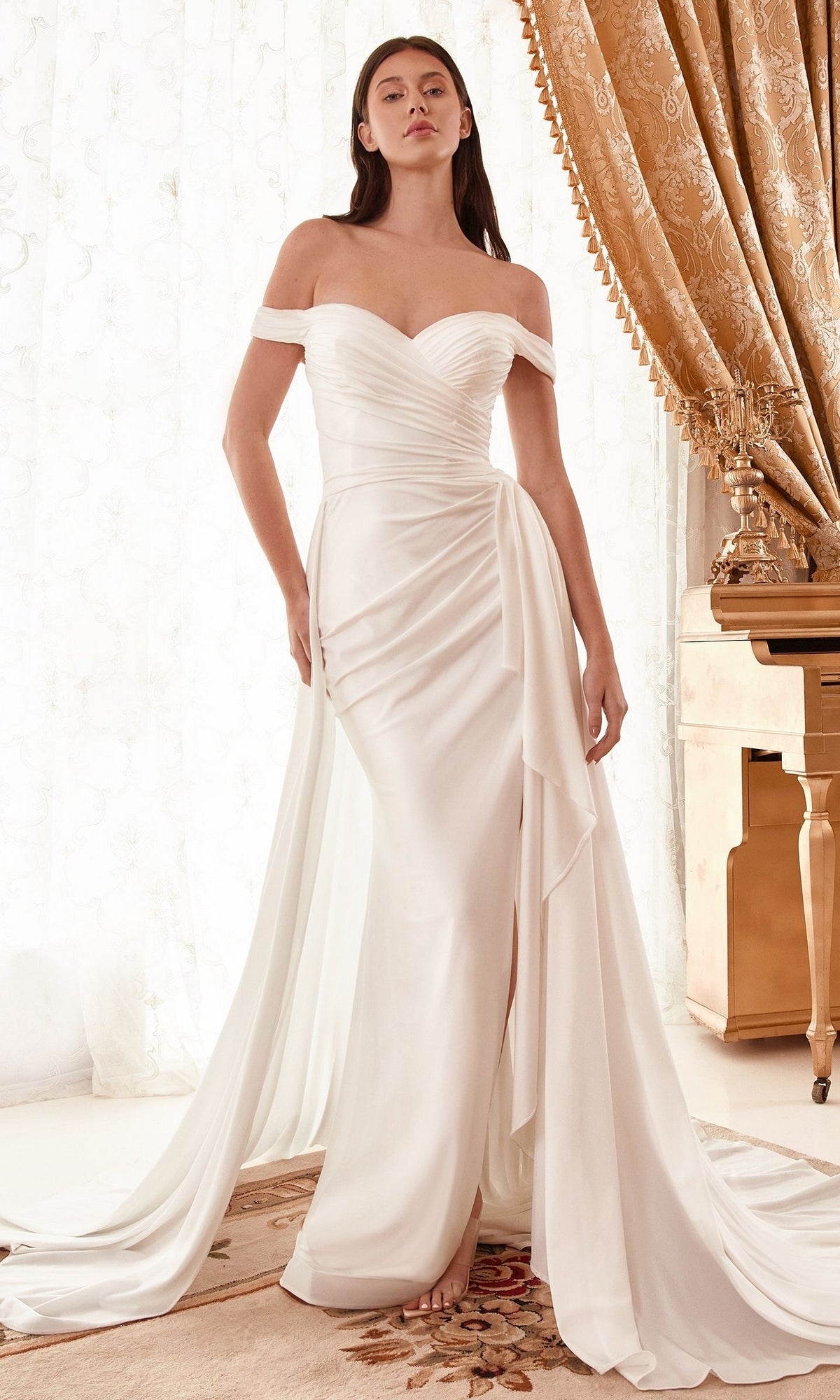 A woman stands indoors next to a gold piano and ornate curtains, wearing the Off-Shoulder Long Off-White Wedding Dress WN315 featuring draped fabric and a long train.
